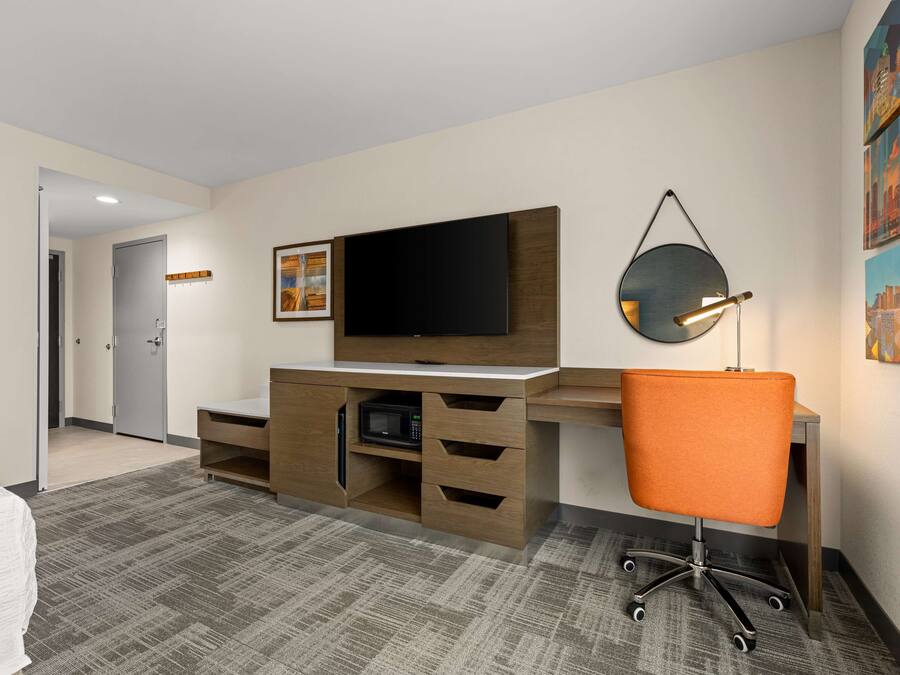 Habitación de hotel con escritorio de madera, televisor de pantalla plana, microondas, silla de oficina color naranja y una obra de arte con un paisaje urbano en la pared.