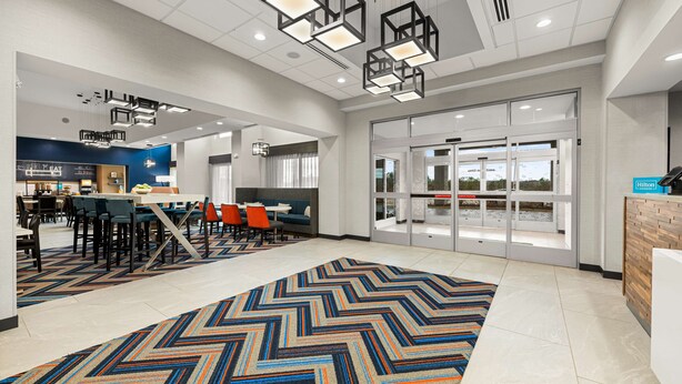A modern hotel lobby with a large, colorful herringbone-patterned carpet, contemporary lighting fixtures, and a seating area with tables and chairs. - Hampton Inn & Suites Bessemer Birmingham (Bessemer)