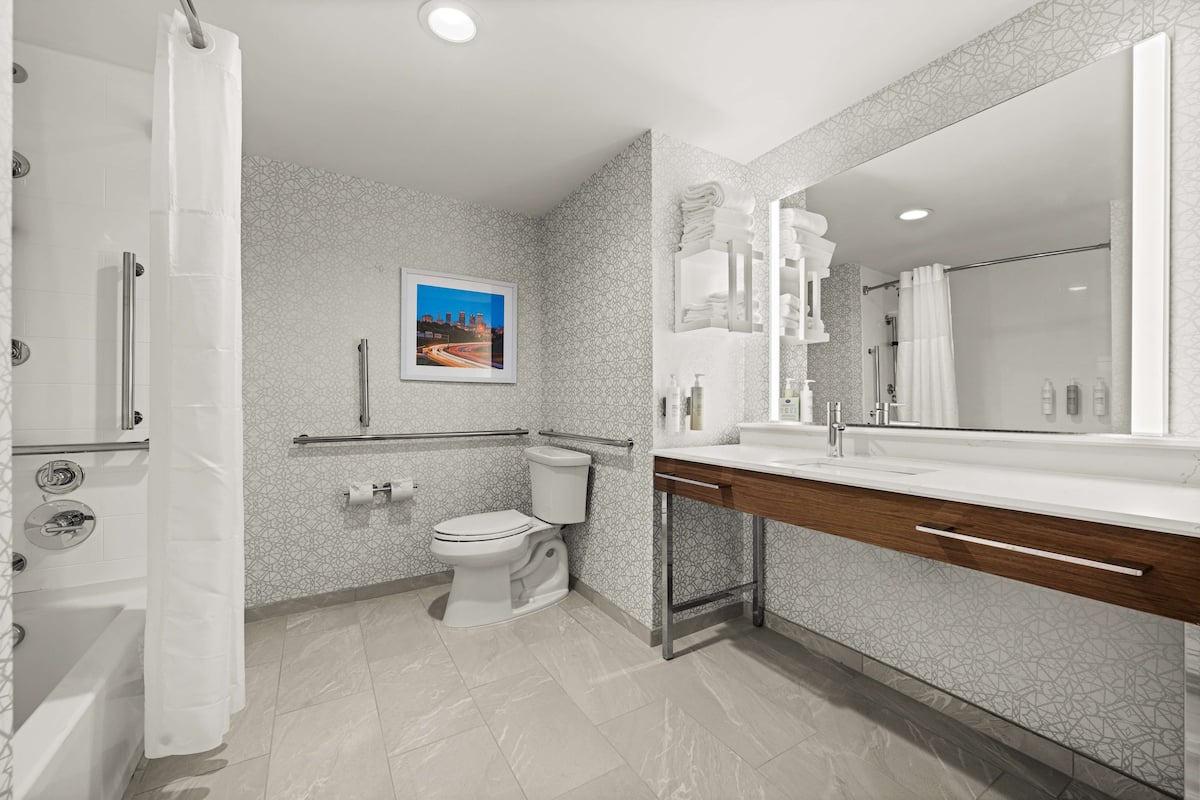 A modern bathroom with a toilet, bidet, and shower. It features a large mirror, a sink with a wooden countertop, and patterned tiles.