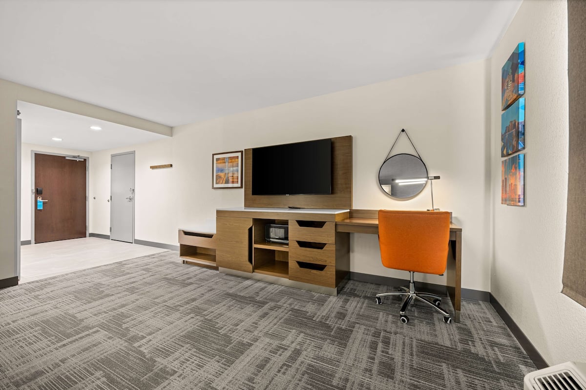 A hotel room with a flat-screen TV, a desk with a chair, a large mirror, and artwork on the wall.