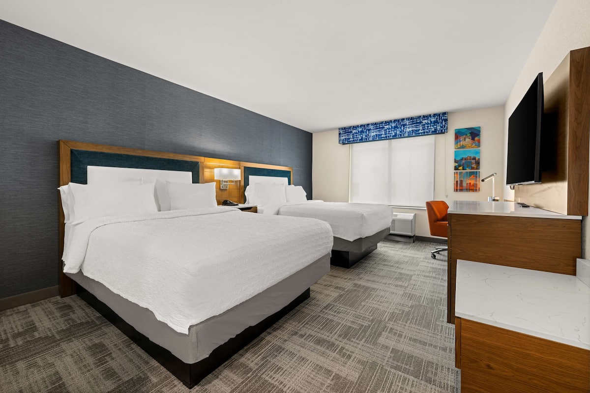 A hotel room with two beds, a desk, a chair, a television, and artwork on the wall.