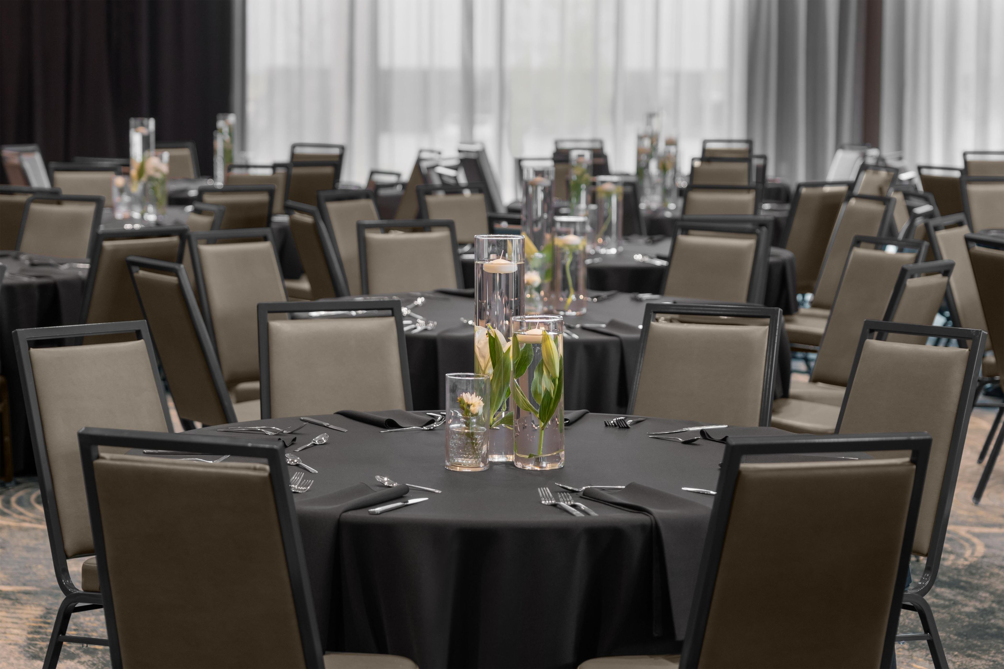 Un salón de conferencias con varias mesas, cada una dispuesta con cubiertos, vasos y jarrones con flores.