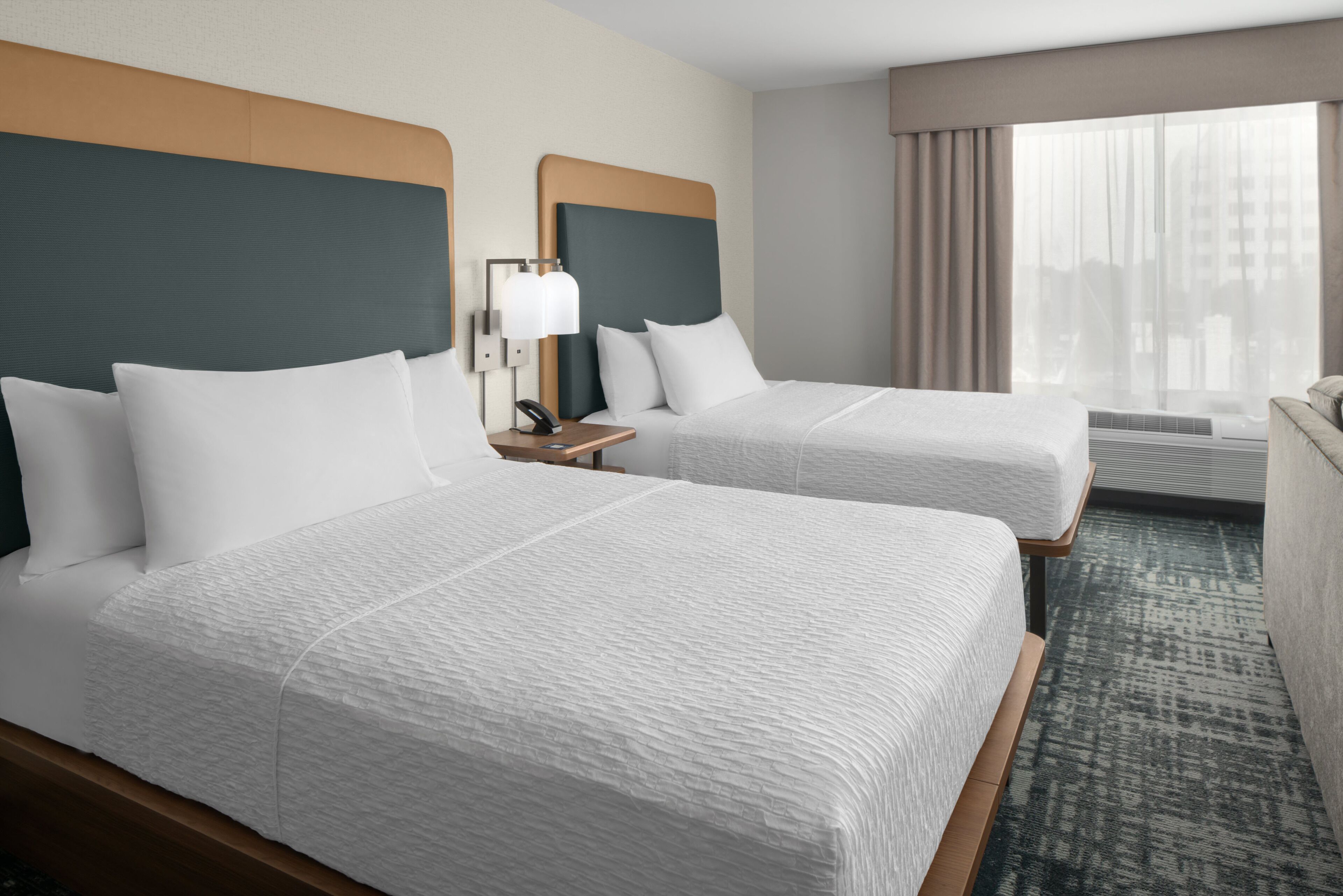 Habitación de hotel con dos camas, una mesita de noche con lámpara, una ventana con cortinas y vista a edificios.