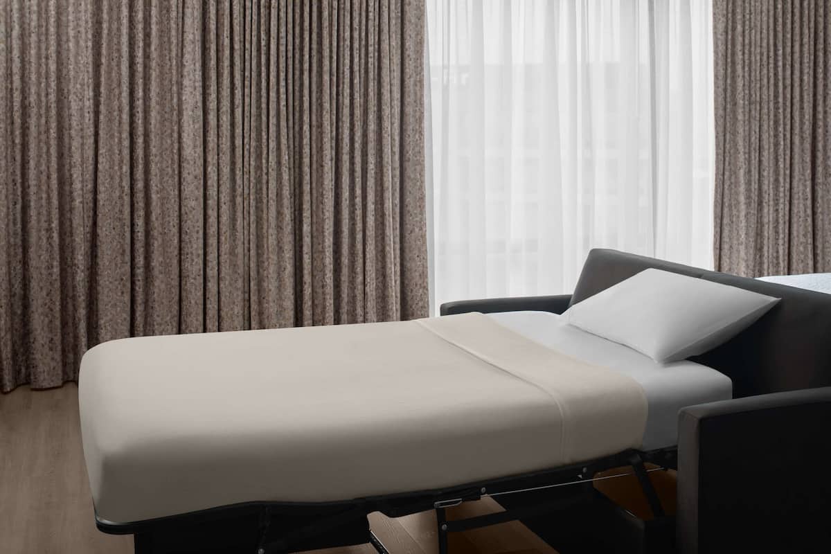 A single bed in a room with a view of a building through sheer curtains.