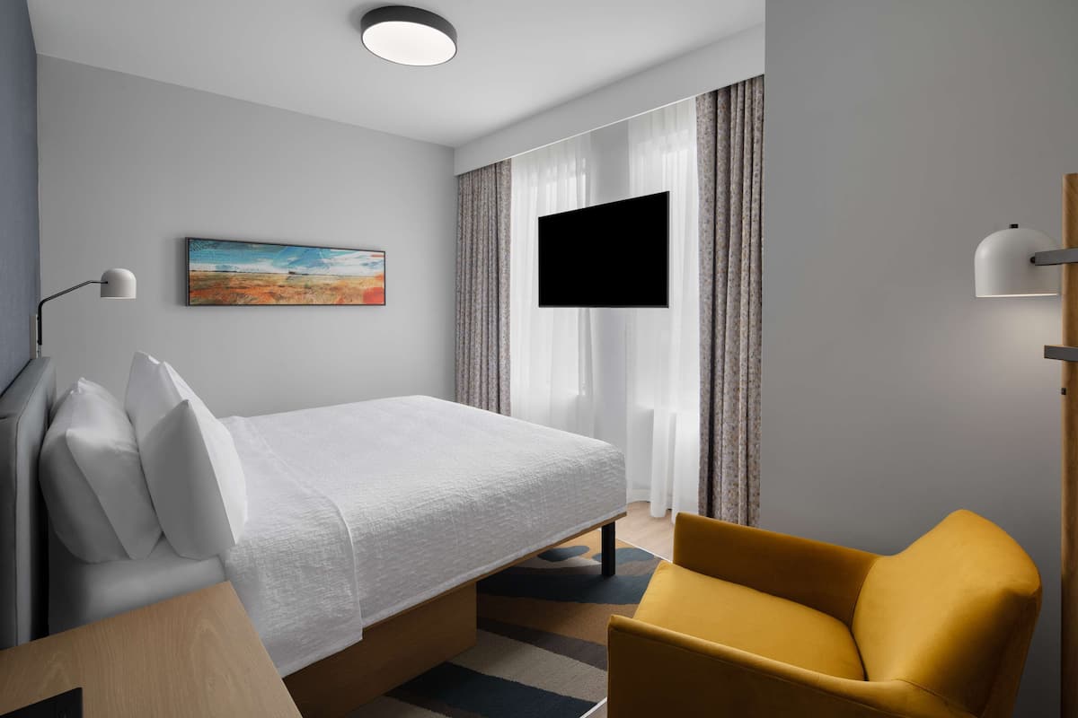A hotel room with a bed, a yellow armchair, a flat-screen TV, a painting on the wall, and a window with curtains.