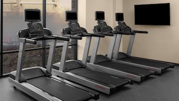 Four Life Fitness treadmills in a gym with large windows and a flat-screen TV on the wall.
