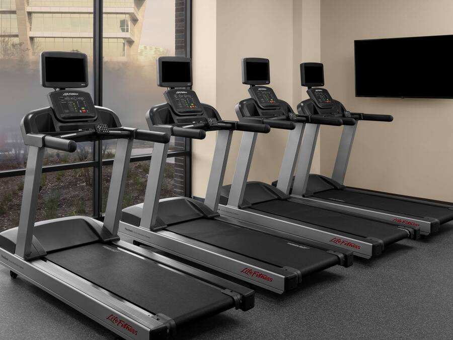 Four Life Fitness treadmills in a gym with large windows and a flat-screen TV on the wall.