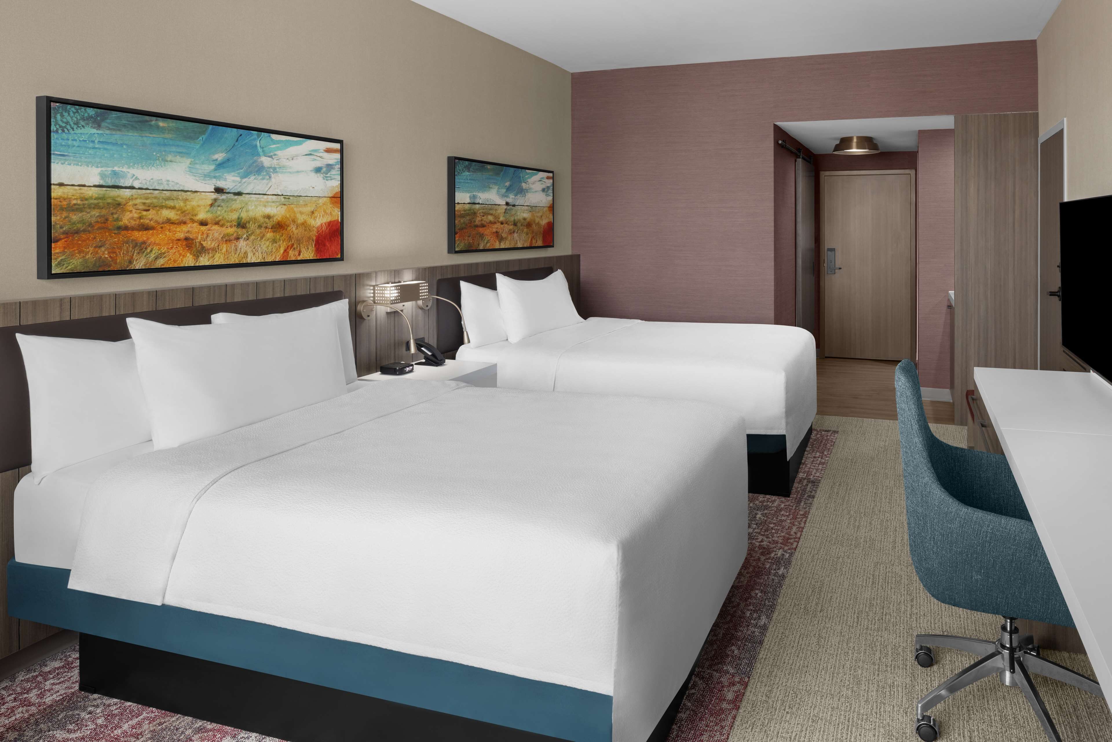 A hotel room with two beds, a desk, a chair, and two paintings on the wall.
