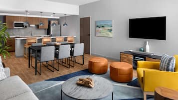 Una sala moderna con un sofá gris, una mesa de centro redonda y un sillón amarillo. Hay una zona de cocina con armarios grises y un televisor de pantalla plana montado en la pared.