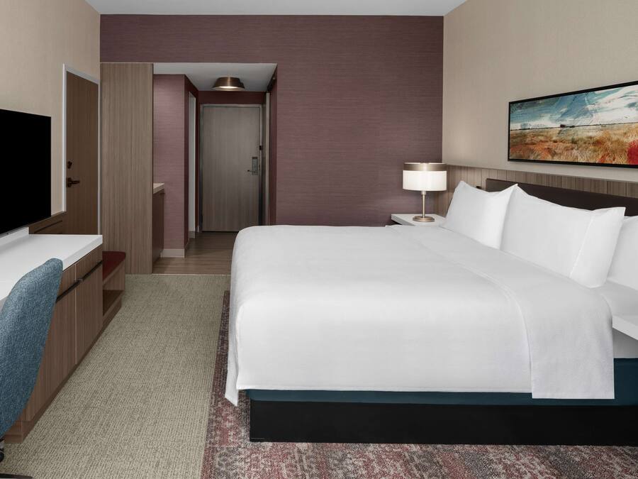 A hotel room with a bed, a desk with a chair, a TV, and a painting on the wall.