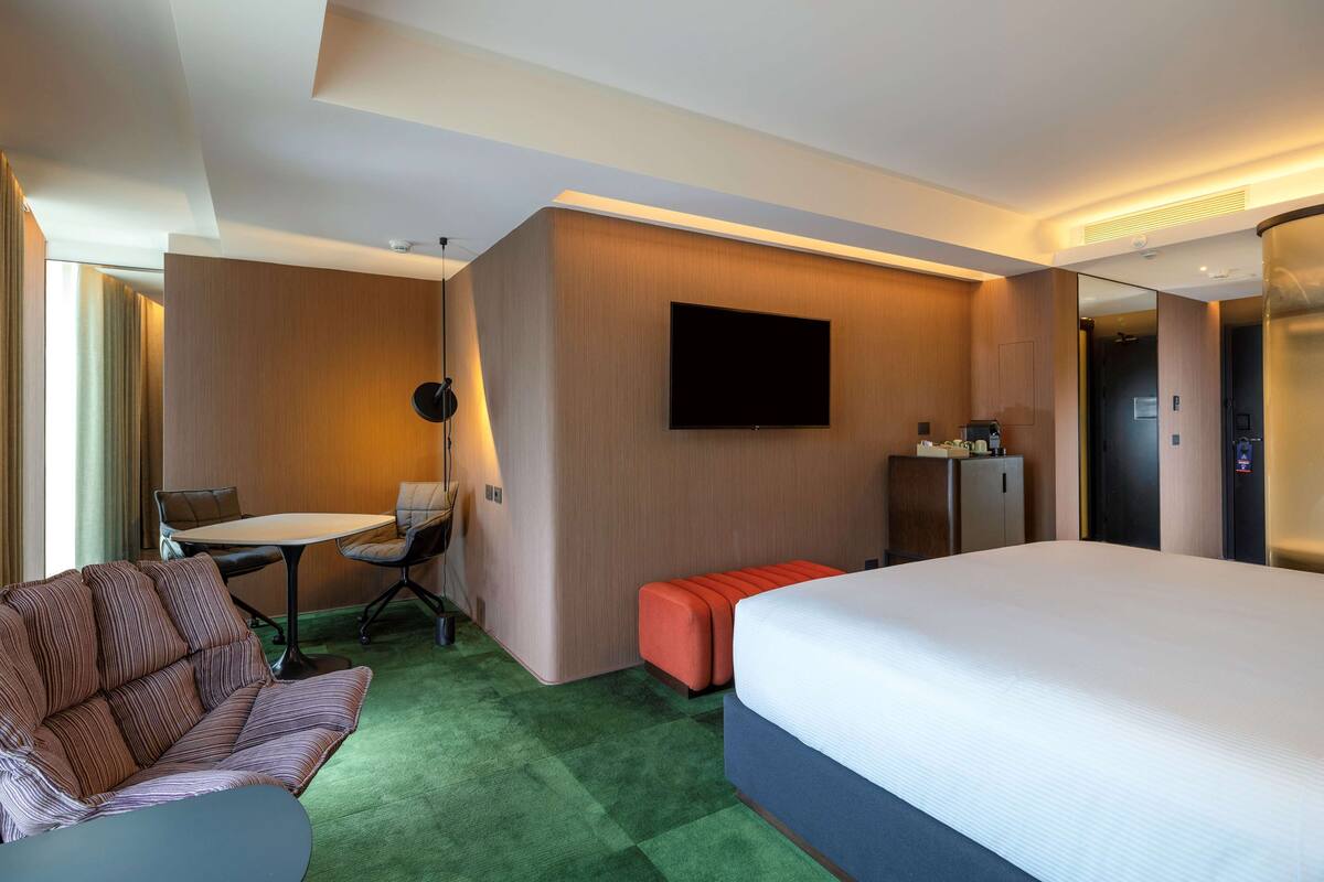 A hotel room with a large bed, a sofa, a desk with a chair, a flat-screen TV mounted on the wall, and a minibar.