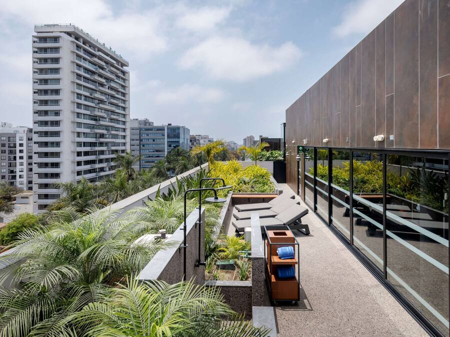A rooftop terrace with a modern design, including a seating area with loungers and a small wooden table, surrounded by lush greenery and a view of the cityscape.
