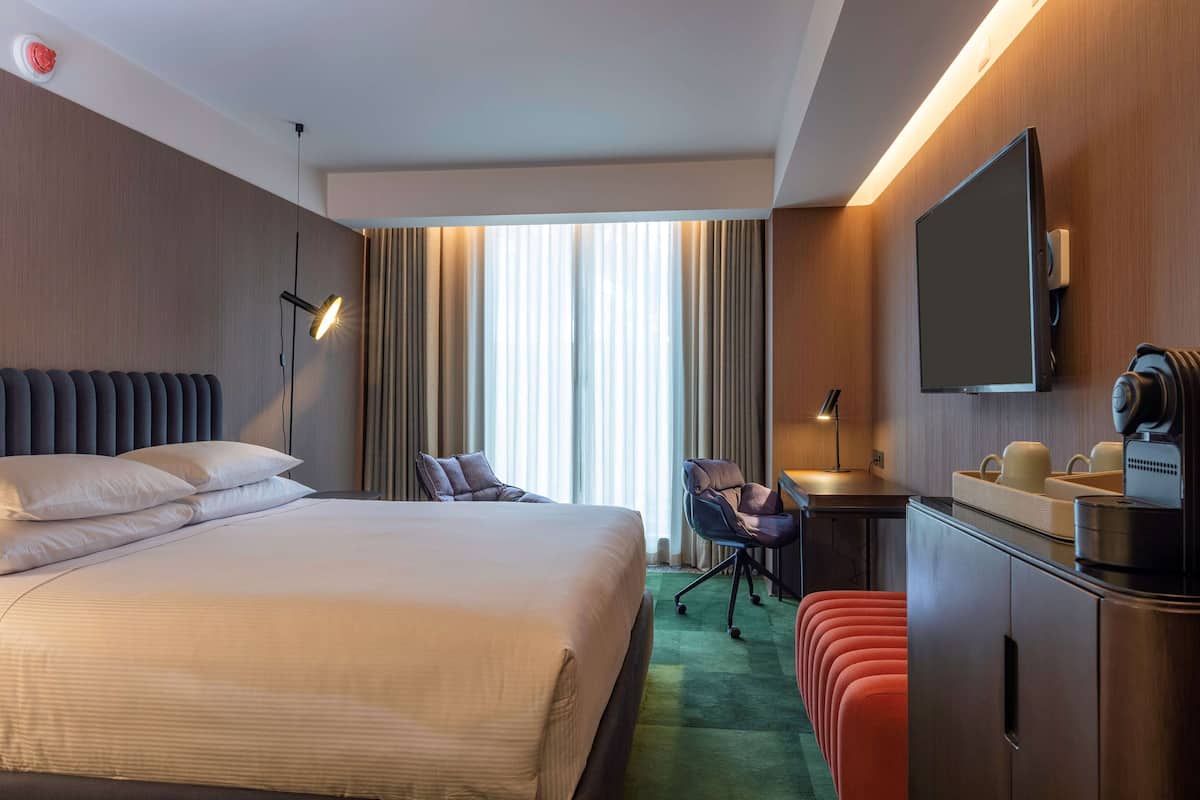A hotel room with a large bed, a desk, a chair, a flat-screen TV, and a minibar.