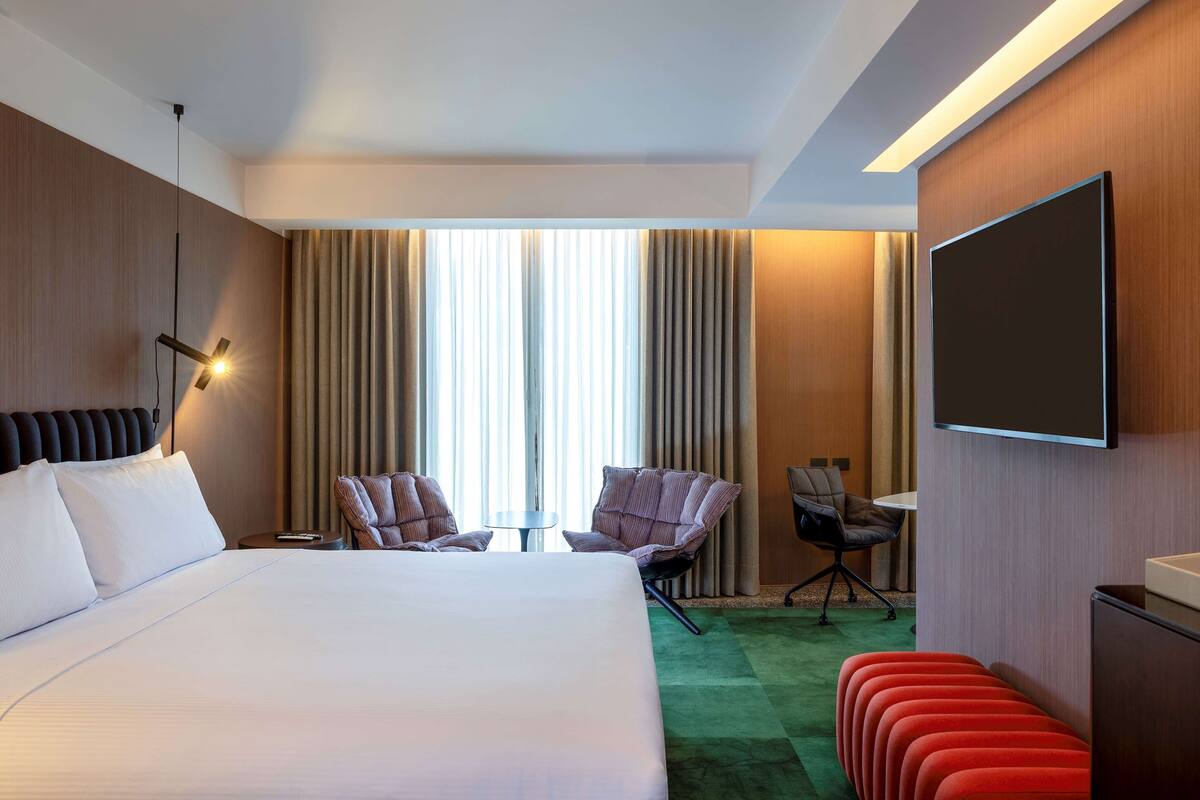 A hotel room with a large bed, two armchairs, a small table, a TV, and a green carpet.