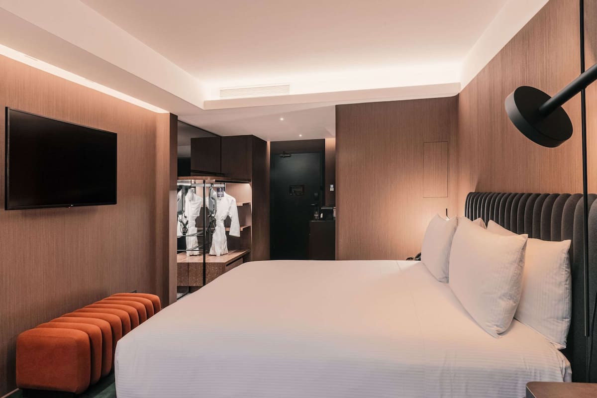A modern hotel room with a large bed, a flat-screen TV mounted on the wall, a walk-in closet, and a seating area with ottomans.