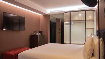 Una habitación de hotel moderna con una cama grande, un televisor de pantalla plana montado en la pared, un pequeño armario de madera y un banco rojo.