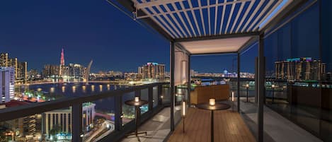 A modern rooftop terrace with a city view, featuring a wooden floor, two tables with candles, and a glass railing.