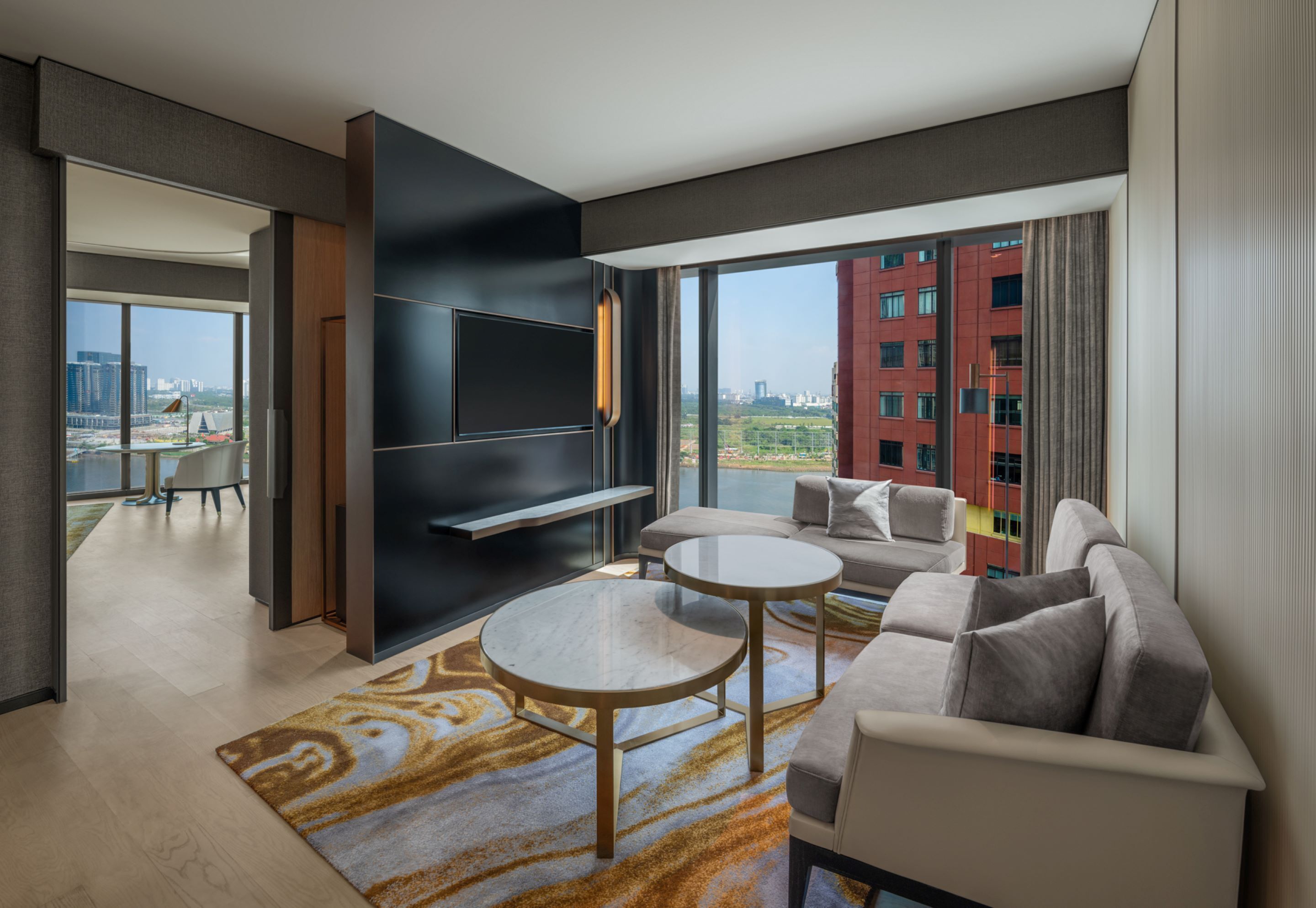 A modern hotel room with a large window, a sofa, two round tables, and a flat-screen TV.