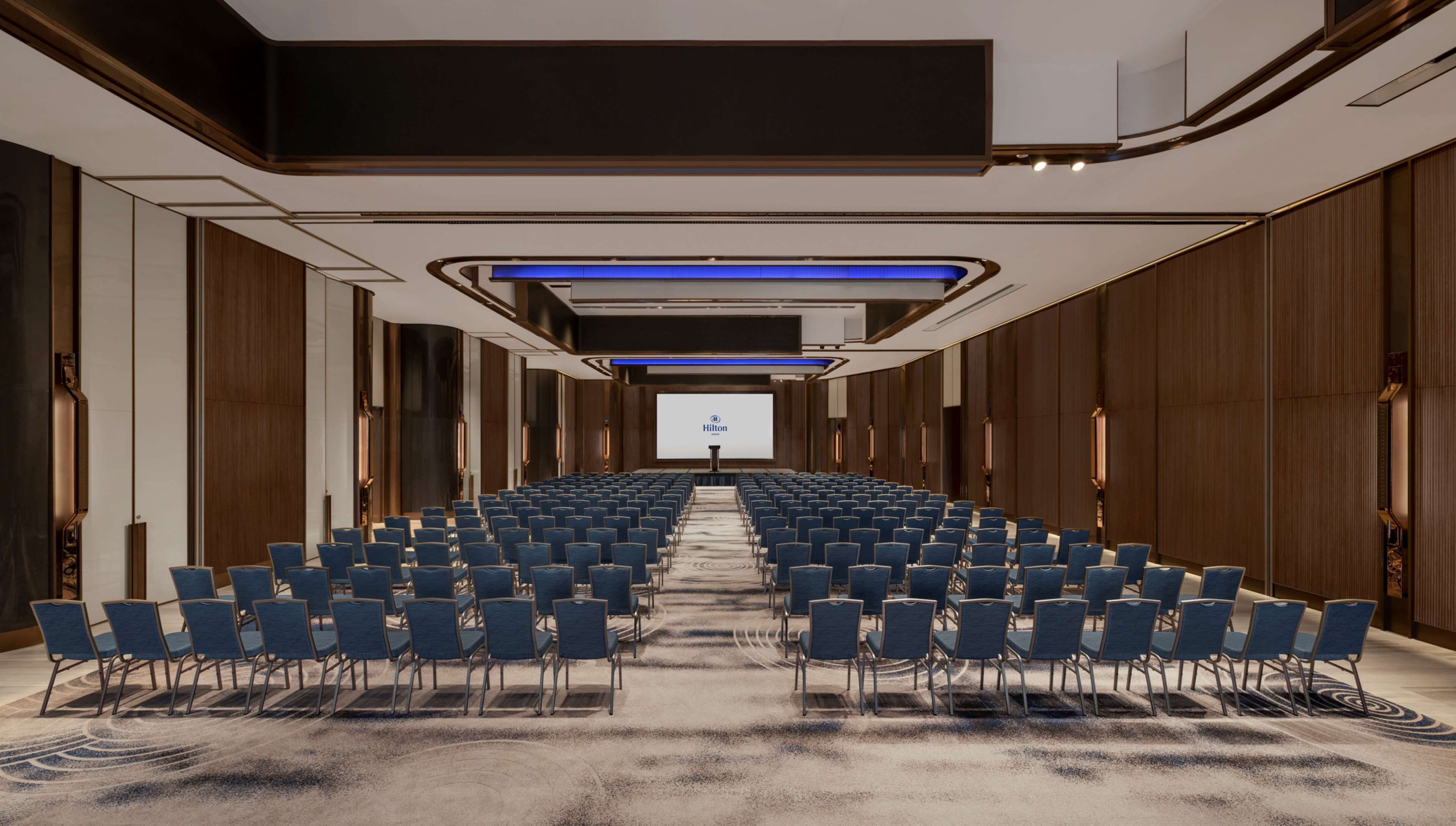 A large, modern conference room with rows of blue chairs, a podium, and a large screen displaying the Hilton logo.