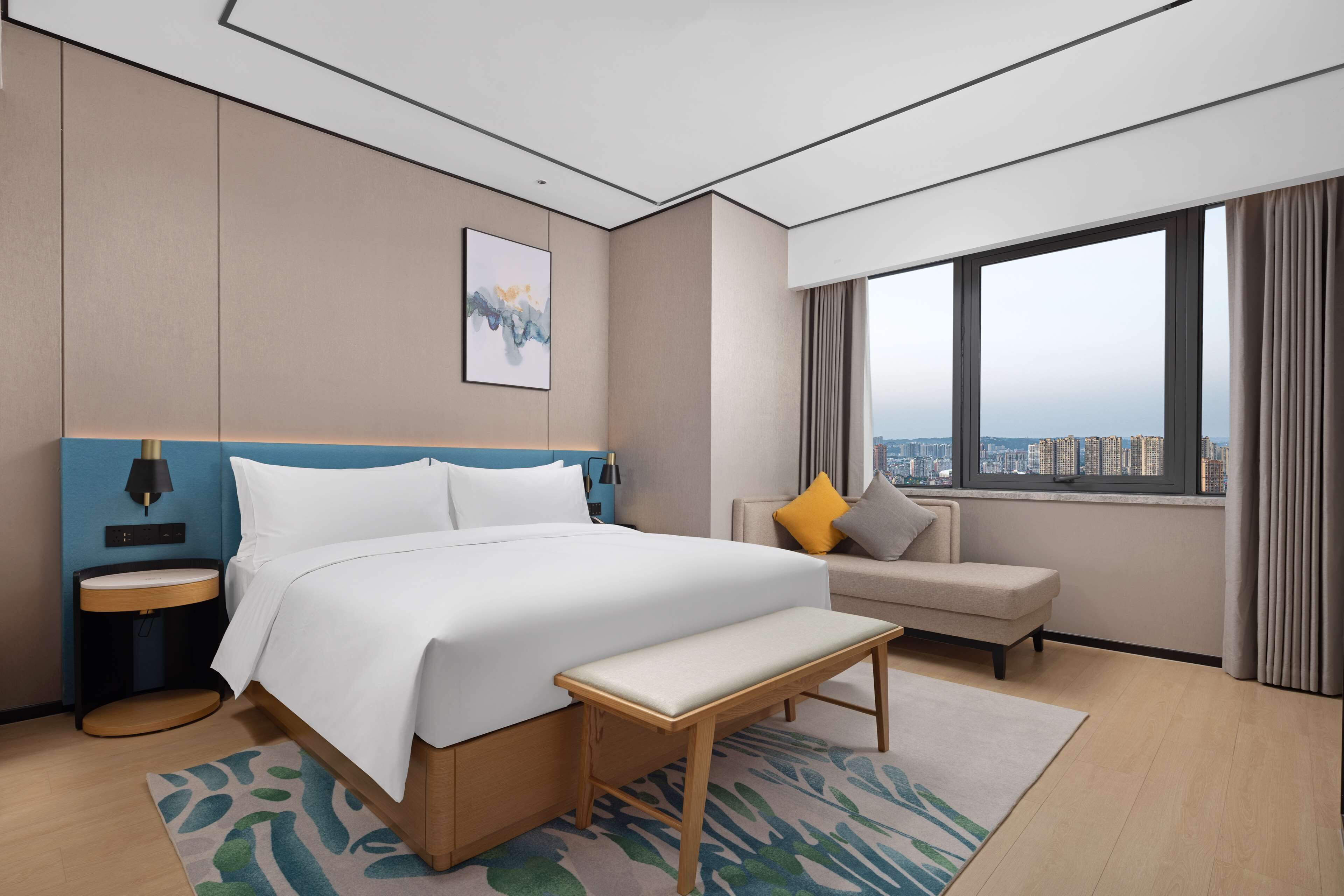 Habitación de hotel moderna con una cama grande, un banco, un sofá, una mesita de noche y vistas a la ciudad a través de grandes ventanales.