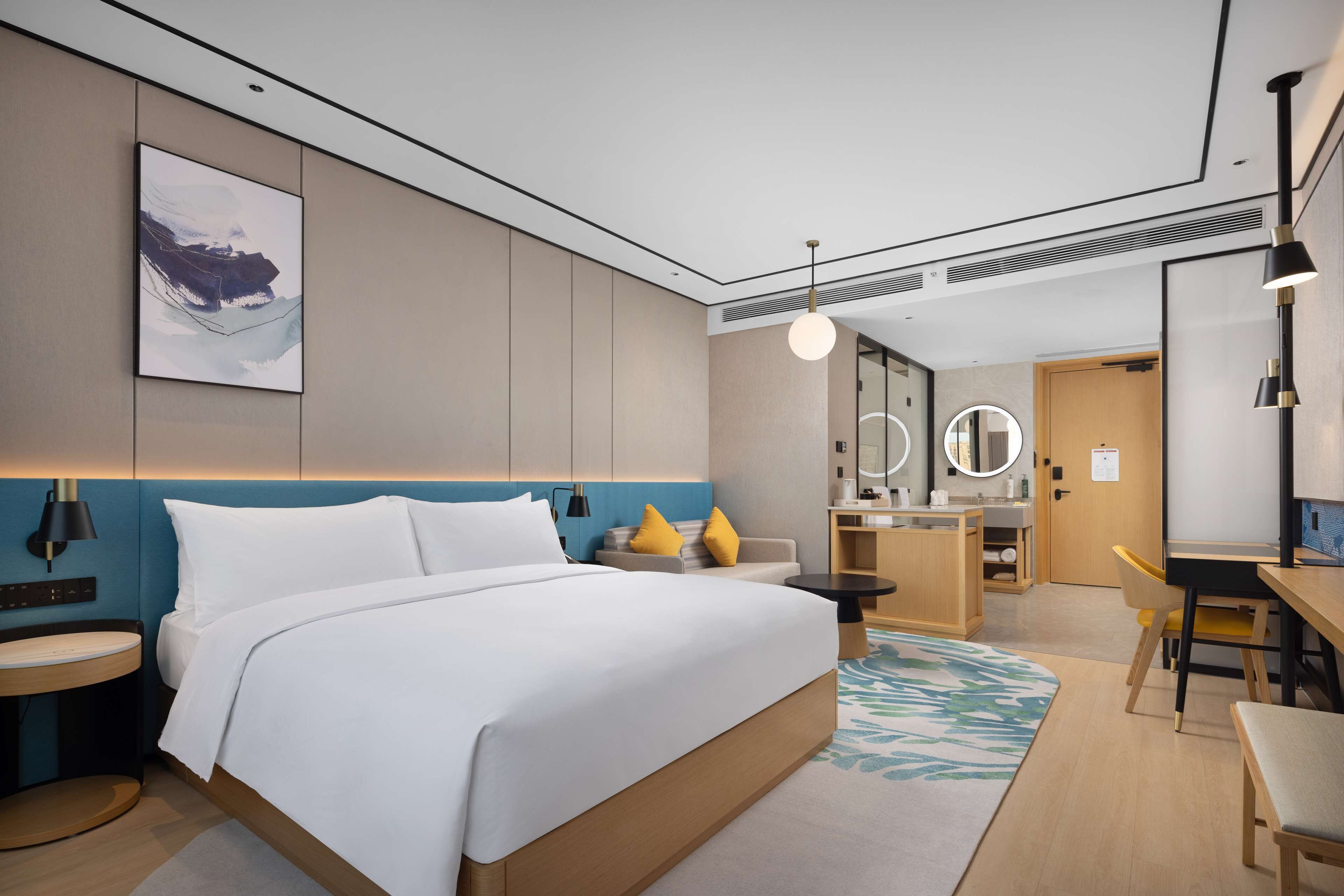 A modern hotel room with a large bed, bedside tables, a desk, and a chair. There is a bathroom with a mirror and a sink.