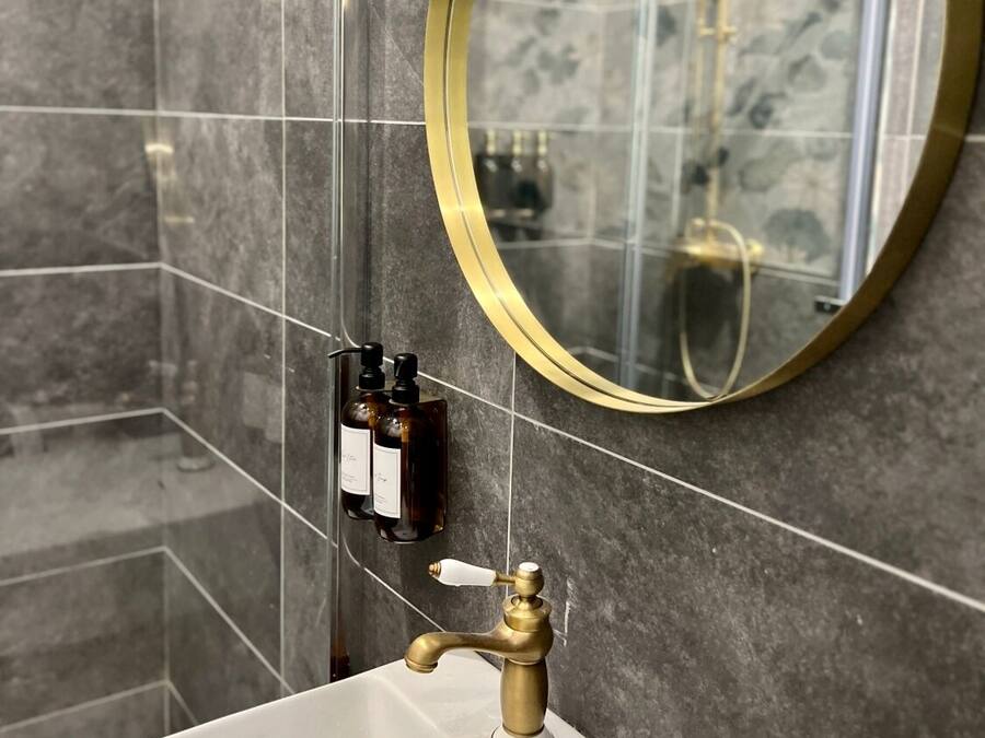 A bathroom with a white sink, gold faucet, and a round mirror with a gold frame. There are two bottles of liquid soap on the wall.