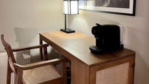Un pequeño escritorio de madera con una cafetera negra, una silla de fibras naturales y un cuadro enmarcado de montañas en la pared.