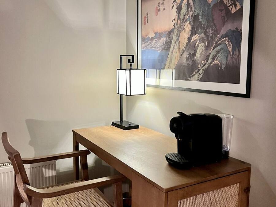 A small wooden desk with a black coffee maker, a woven chair, and a framed painting of mountains on the wall.
