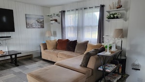 Una sala de estar con un sofá color beige, una mesa de centro y un televisor. Hay objetos decorativos en estanterías y una alfombra en el suelo.