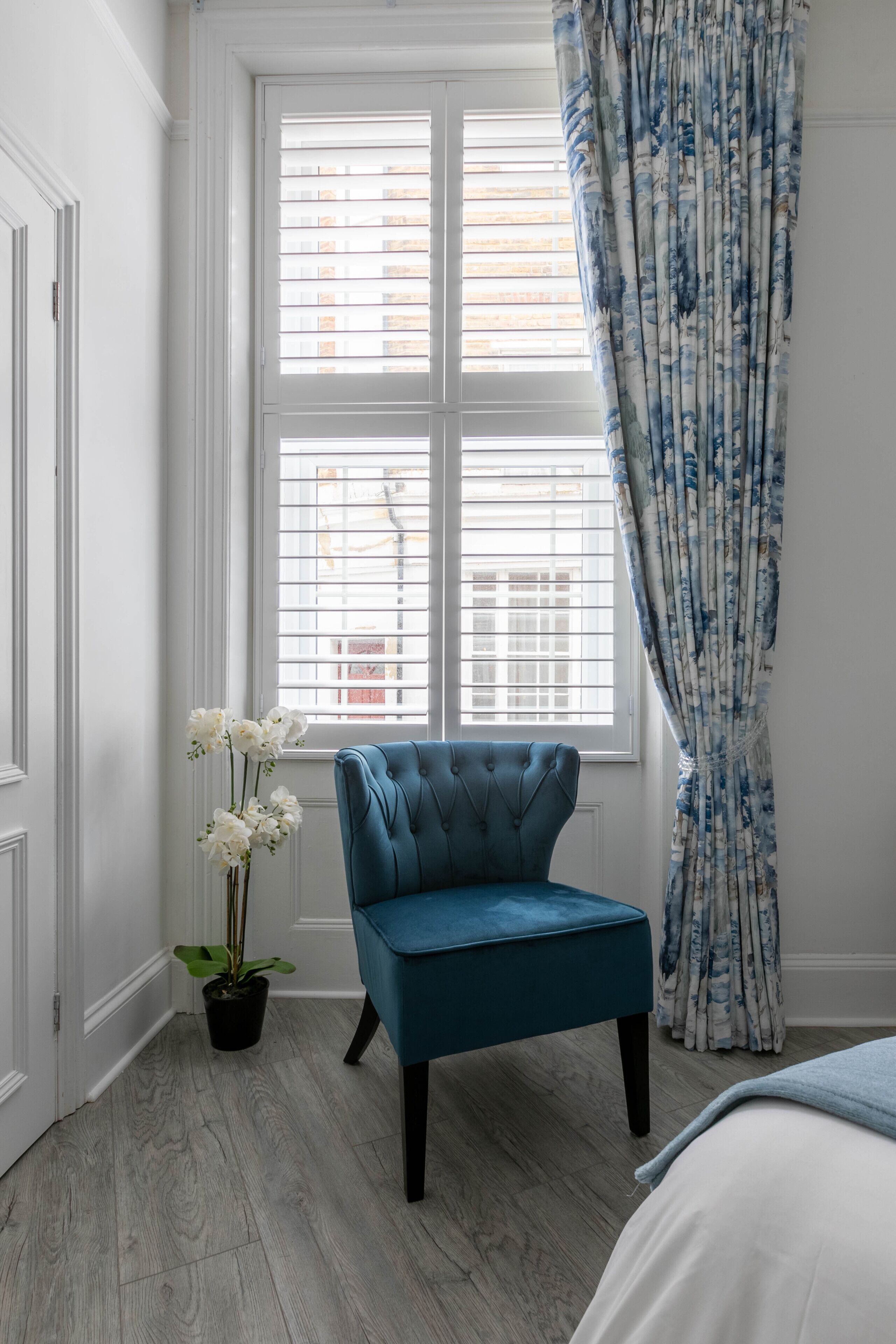 A room with a large window, a blue chair, a potted plant, and a bed.