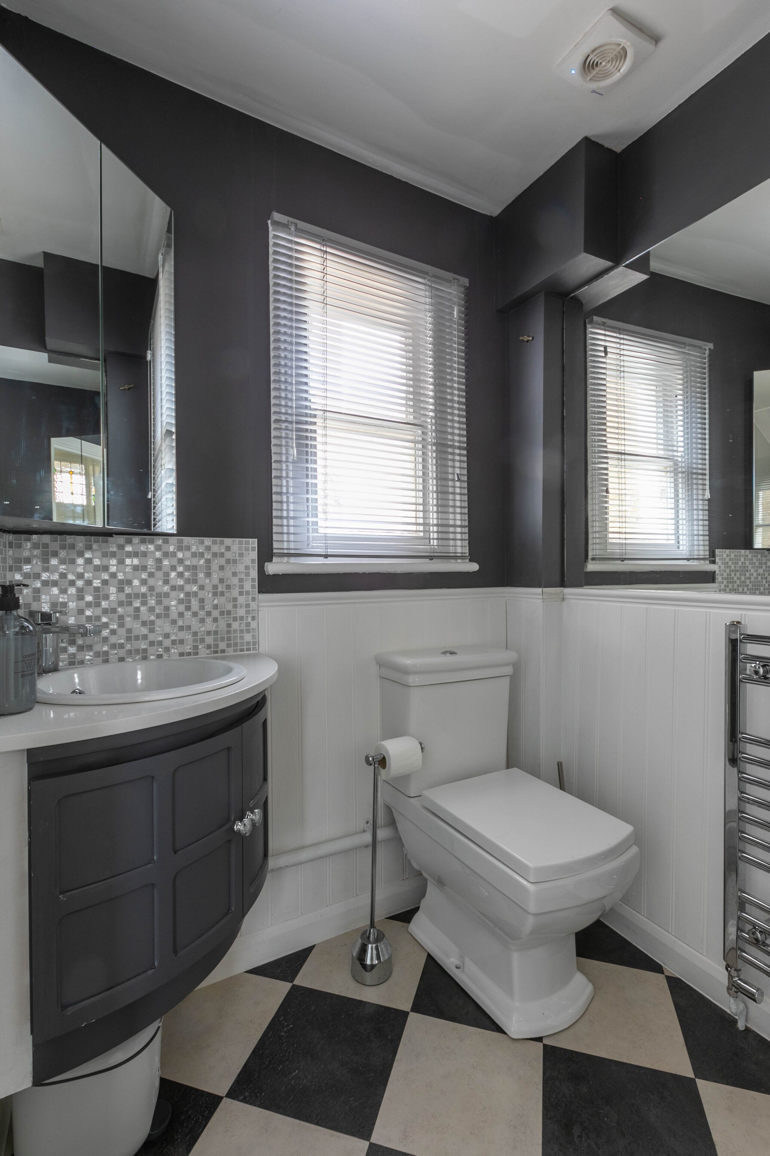 Une salle de bain avec des toilettes blanches, un lavabo et un miroir. Il y a des fenêtres avec des stores et un pare-feu en carrelage.