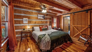 A wooden cabin bedroom with a bed, a nightstand, a lamp, a ceiling fan, and a view of the outdoors. - Epic Retreat I Pickleball I Basketball I Arcade I Putting Green I Air Hockey (Sevierville)
