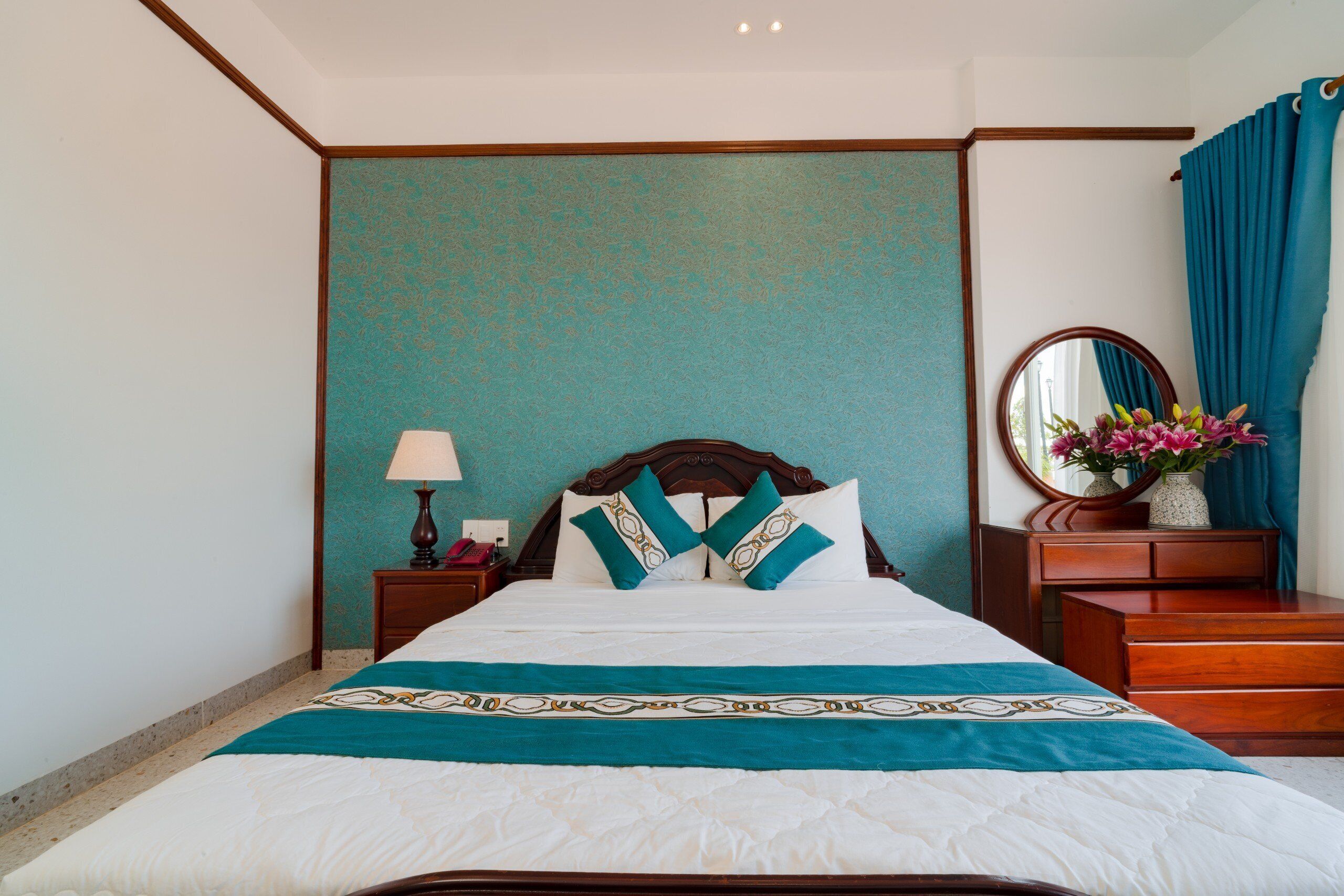 A neatly made bed with a teal bedspread and pillows, a wooden nightstand with a lamp, a dresser, and a mirror with a vase of flowers.