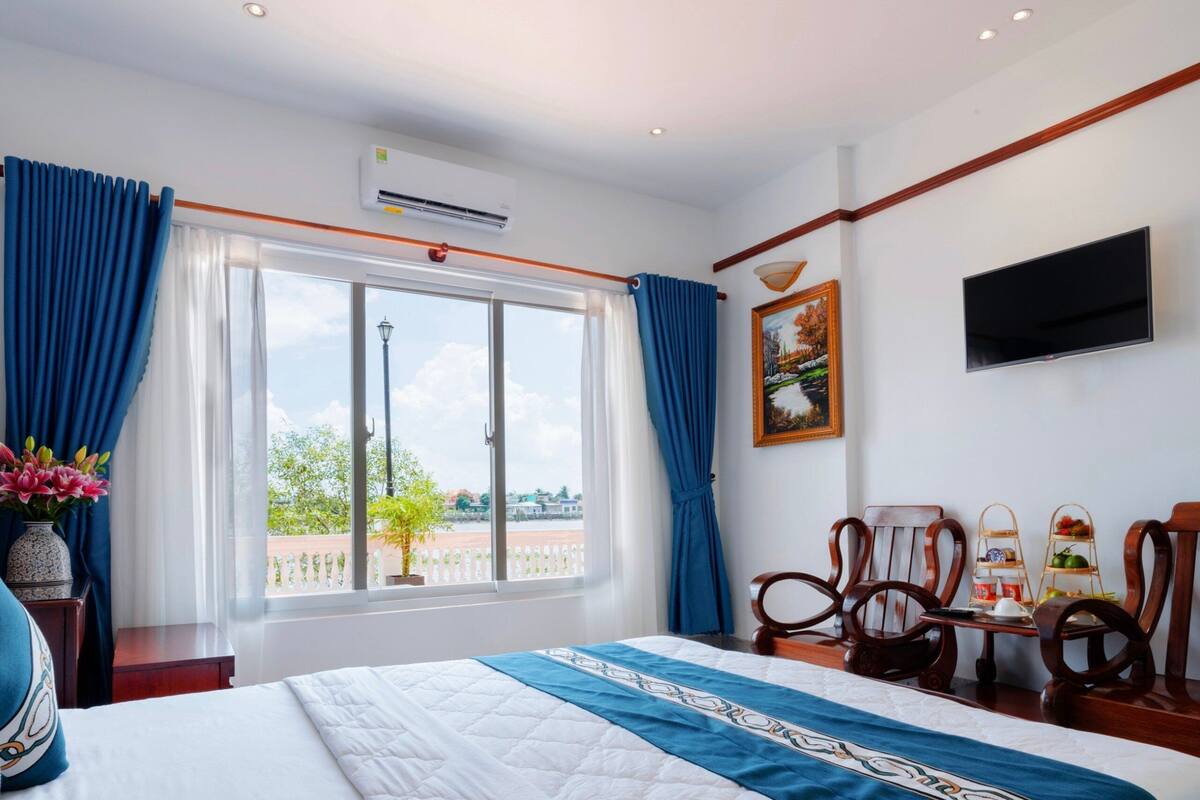 Ein Hotelzimmer mit einem großen Bett, einem an der Wand montierten Fernseher und Blick ins Freie durch ein großes Fenster.
