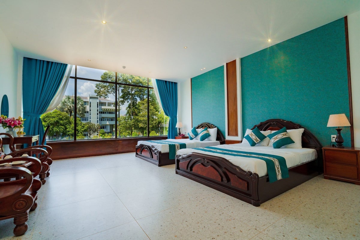 Ein Hotelzimmer mit zwei Betten, einem großen Fenster mit Vorhängen, Blick auf Grünflächen und einem gefliesten Boden.