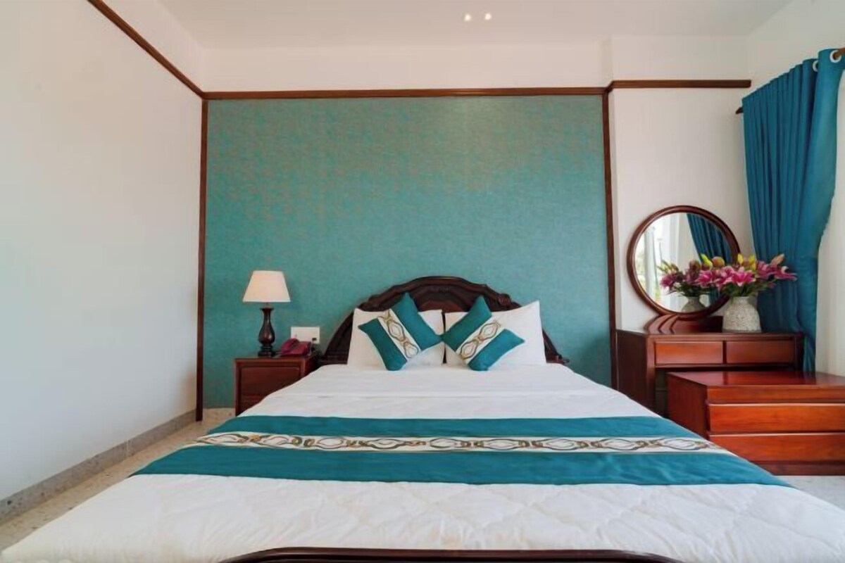 A neatly made bed with a patterned bedspread, a wooden headboard, and a nightstand with a lamp and a vase of flowers.