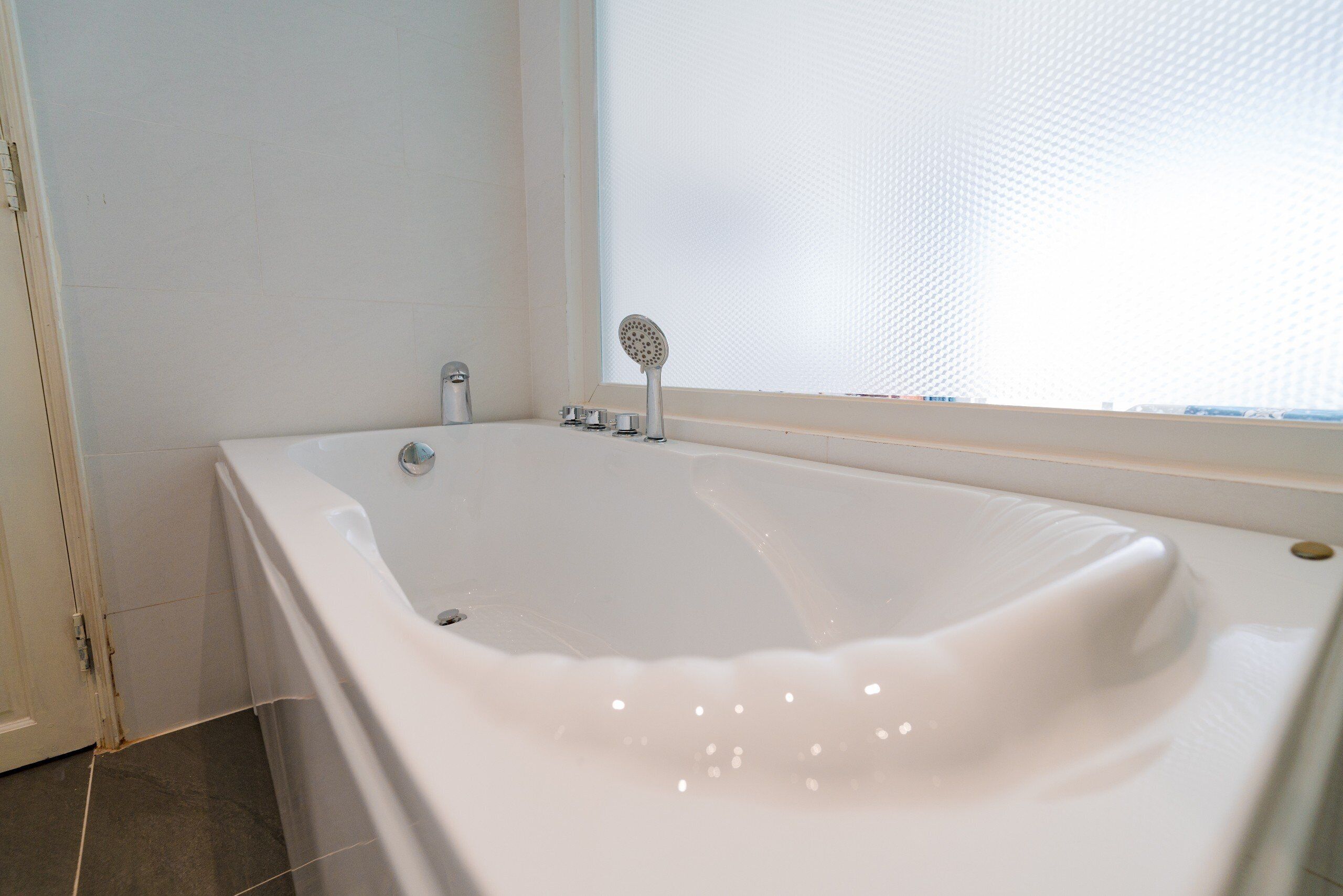 Een moderne badkamer met een groot bad, een douchekop en een raam met gezandstraald glas.