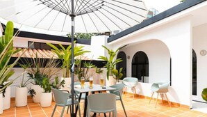 Eine Terrasse im Freien mit Tisch und Stühlen, umgeben von Topfpflanzen und einem großen Sonnenschirm.