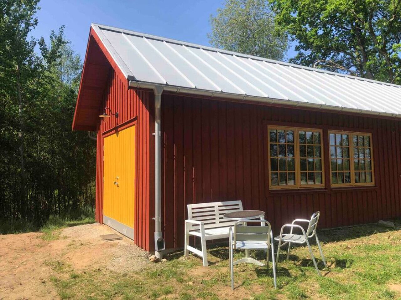 En röd byggnad med ett tak av metall, en gul dörr och ett fönster med ett rutmönster. Utanför finns det två vita stolar och ett bord.