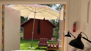 Ein Schlafzimmer mit einem Bett, einem Nachttisch, einer Lampe und Blick auf einen roten Schuppen und Liegestühle draußen.