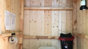 Ein kleines Badezimmer aus Holz mit Toilette, einem Holzschemel, einer schwarzen Mülltonne und einer weißen Leuchte.