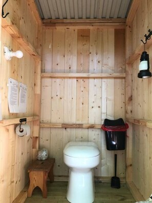 Un baño pequeño de madera con un inodoro, un taburete de madera, un bote de basura negro y una lámpara blanca.
