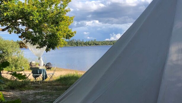 Ein Zelt in der Nähe eines Sees, mit Tisch und Stühlen, umgeben von Bäumen.