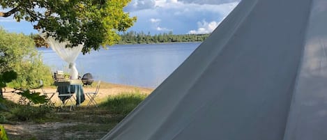 Une tente installée près d’un lac, avec une table et des chaises, le tout entouré d’arbres.