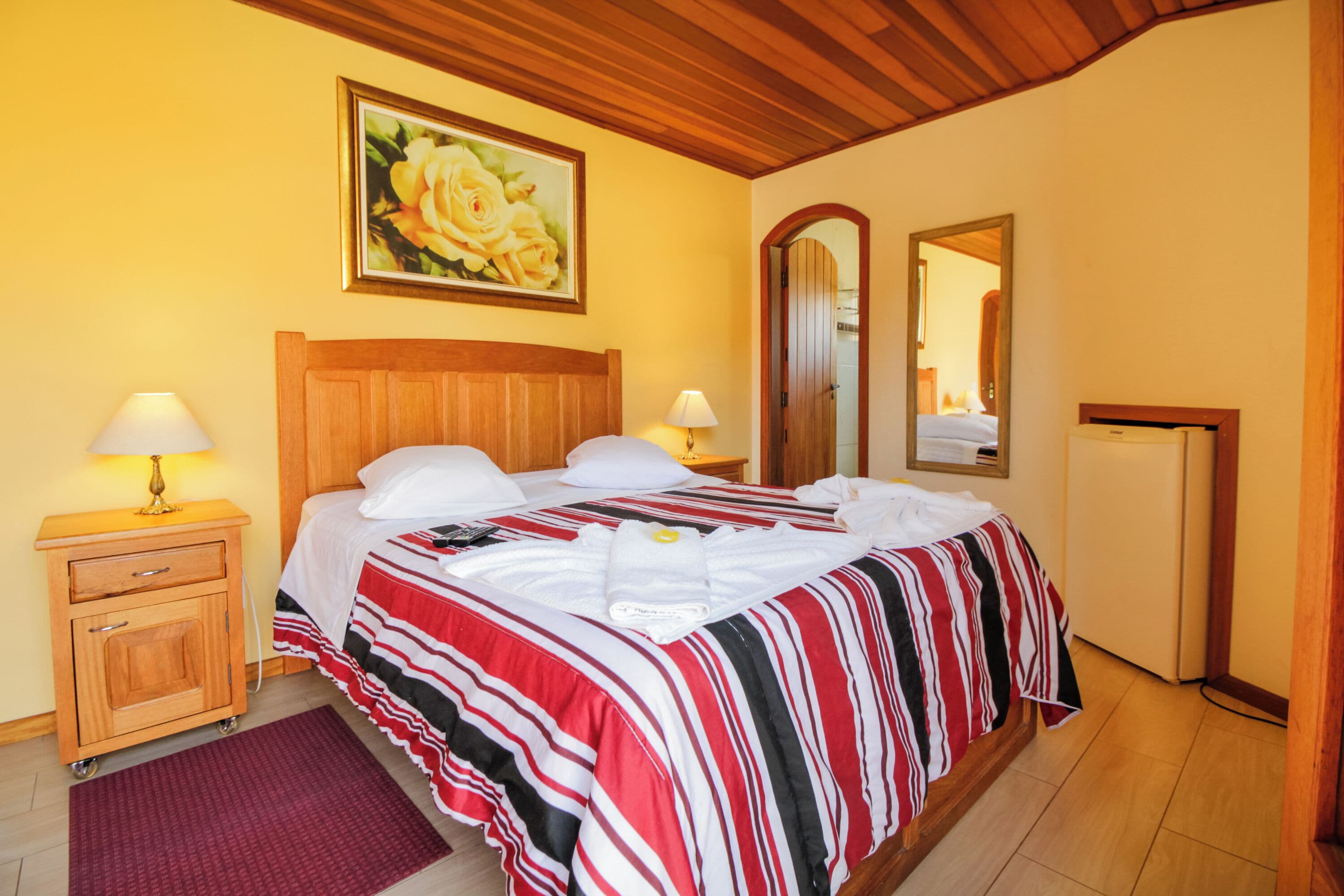 Une chambre d’hôtel avec une tête de lit en bois, un lit recouvert d’une couvre-lit à rayures, une table de chevet avec une lampe, un tapis rouge, un tableau représentant des roses et un miroir.