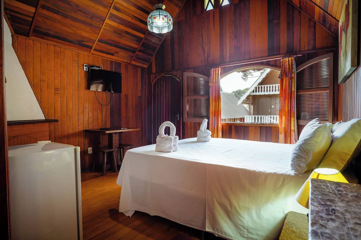 Ein Zimmer in einer Holzhütte mit einem Bett, einem kleinen Tisch, einem Kühlschrank und einem Fenster mit Vorhängen.