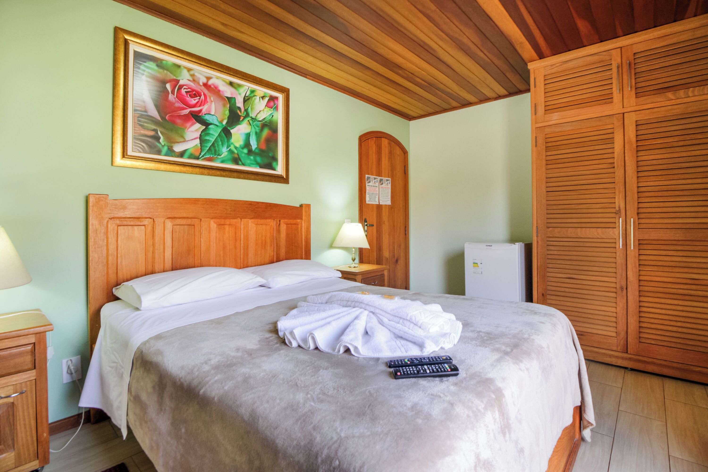 Un letto rifatto con coperta e telecomandi, una testiera in legno, un comodino con una lampada, un quadro con rose e un armadio.