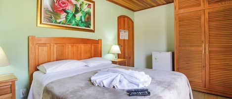 Un letto rifatto con coperta e telecomandi, una testiera in legno, un comodino con una lampada, un quadro con rose e un armadio.