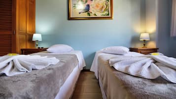 Two single beds with white comforters and towels, a wardrobe, nightstand, and lamp, and a framed floral painting adorn this cozy space.