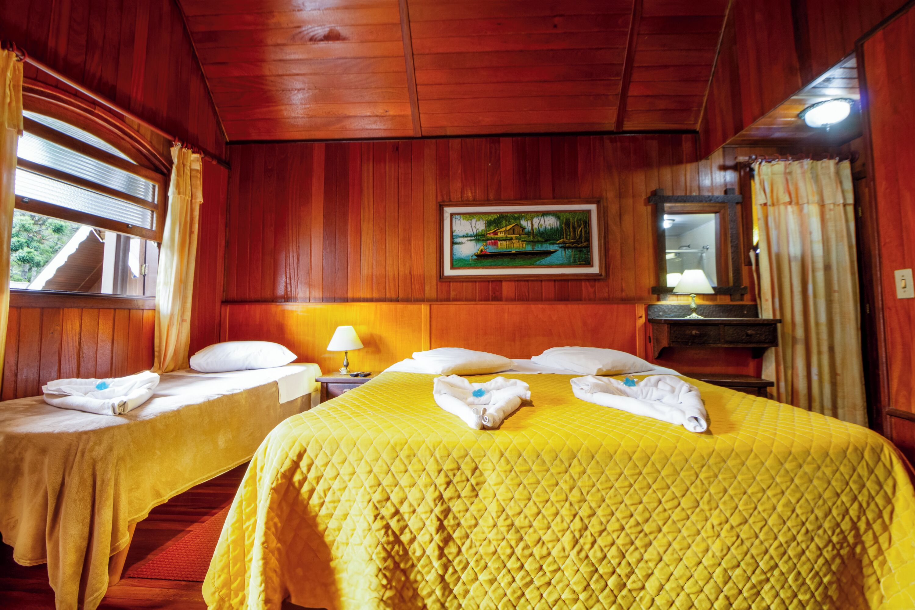 Habitación de hotel con dos camas, paredes de madera, un cuadro, un espejo y una ventana con cortinas.